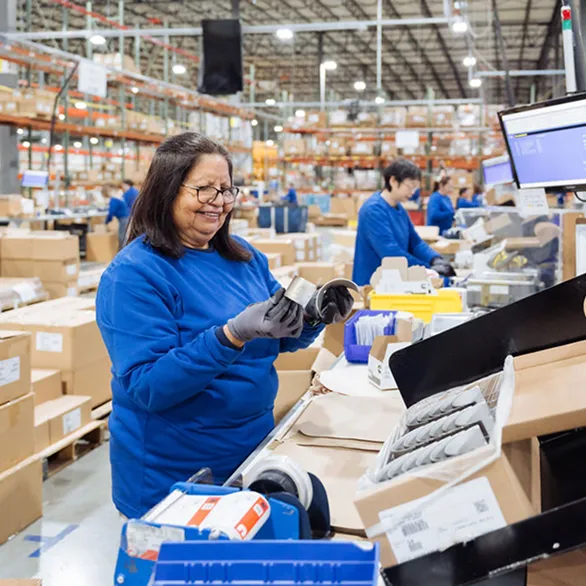 Worker wearing safety glasses feeds material into a machine with rollers while holding a sheet of packaging in a warehouse.