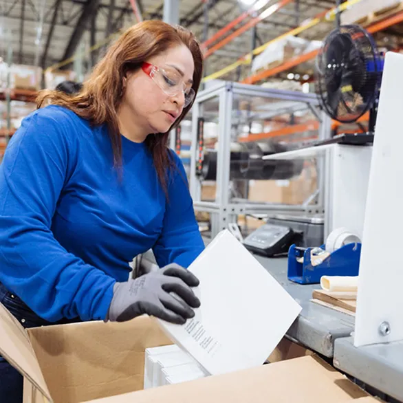 Worker in gloves placing white boxed products into an open cardboard shipping carton at a packing workstation.