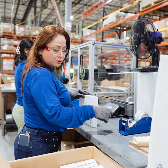 Worker in safety glasses using a handheld scanner to process boxed units at a packing station with conveyor and shelving in background.