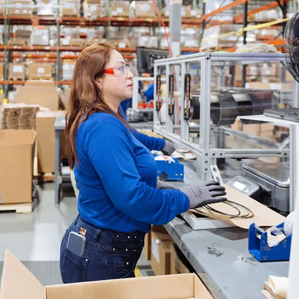 Worker in safety glasses standing at a coil processing station with enclosed rollers, fan, and control interface on a factory floor.
