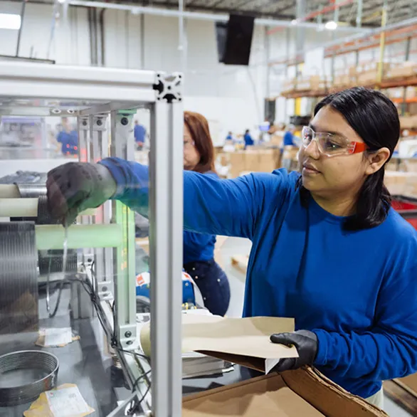 Worker in blue shirt loading sheet material into an enclosed machine with rotating coils and control panel on assembly floor.