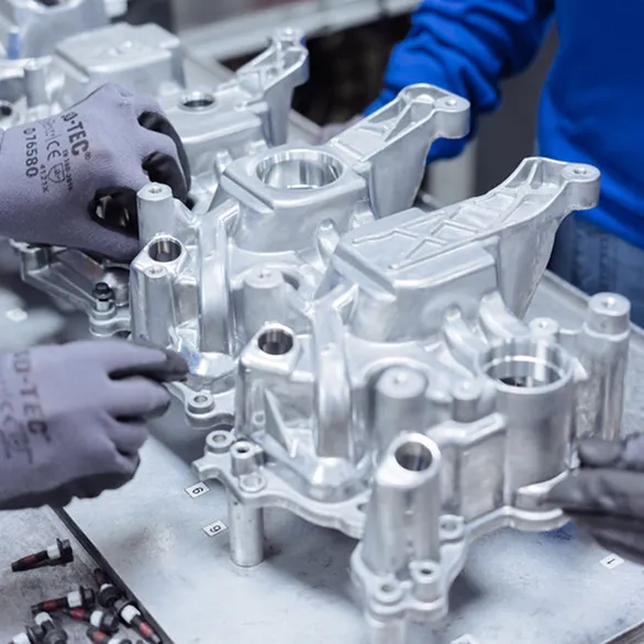 Close view of two pairs of gloved hands positioning a metal casting on a fixture with bolts and alignment pins visible.