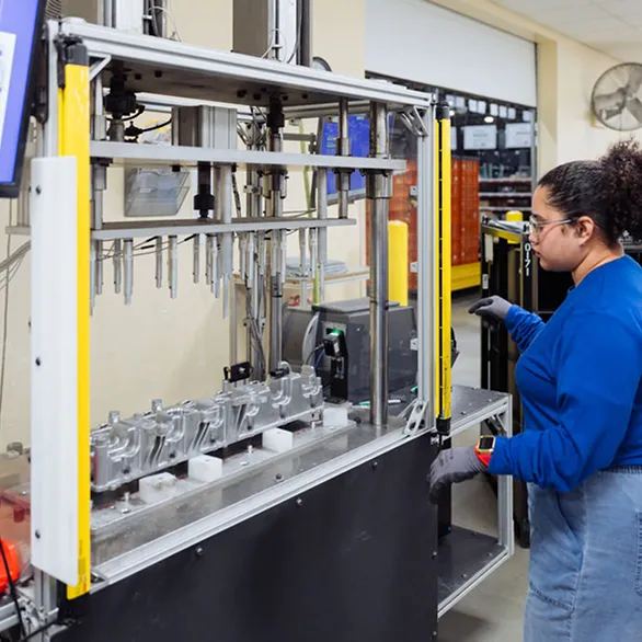 Worker stands at an enclosed fixture station with multiple vertical tools positioned above a metal casting on the work surface.