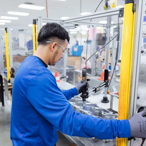 Worker uses a handheld electric screwdriver to install fasteners into a metal casting inside a guarded workstation.