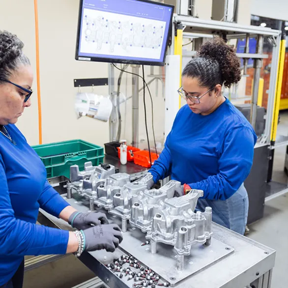 Two workers place fasteners into a metal casting on a fixture with bins of bolts and tools arranged on the work surface.