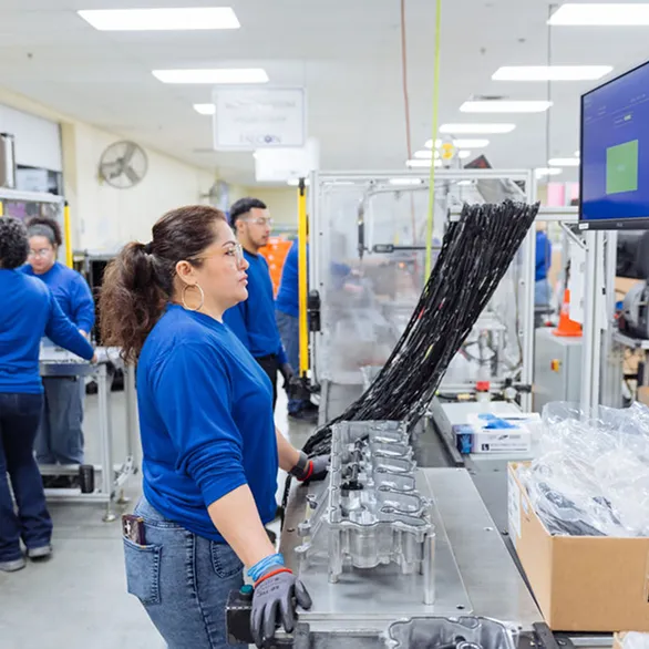 Worker positions a wiring harness over a large metal casting at a workstation with a monitor and plastic bins on the table.