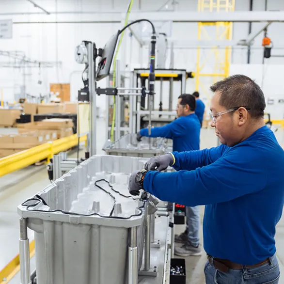 Worker in blue shirt securing wiring inside a metal housing on a conveyor assembly line.