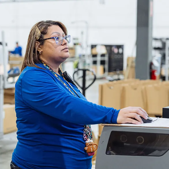 Worker operates a control interface at a testing machine while standing beside boxed components on the production floor.