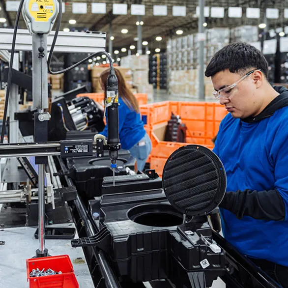 Worker holds a circular cover while positioning it over an opening in a black plastic housing at a workstation.
