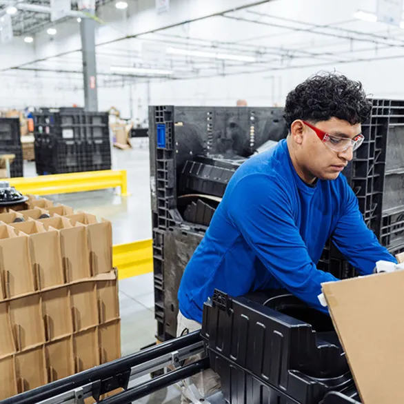 Worker loads or removes a black plastic component from a crate beside a production conveyor.
