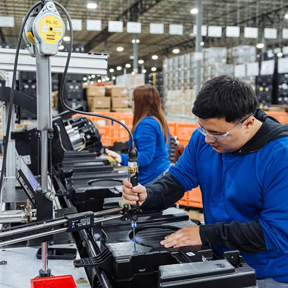Worker uses a handheld fastening tool to secure a component on a black plastic assembly at a testing station.