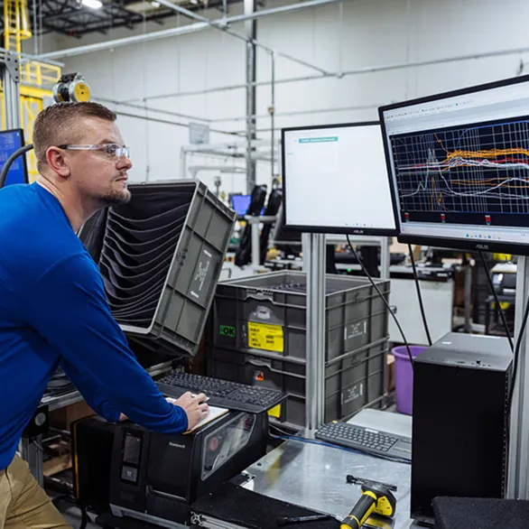 Worker stands at computer workstation reviewing performance graphs displayed on dual monitors in a manufacturing environment.