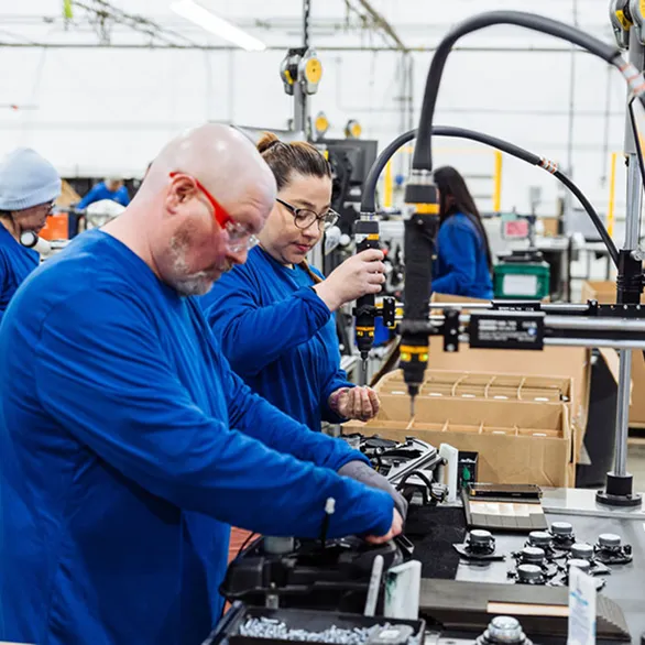 Workers use suspended torque tools to fasten components onto assemblies along a manufacturing line with bins and fixtures.