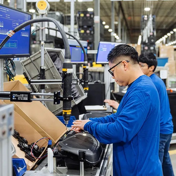 Two workers operate powered tools while assembling black plastic housings at adjacent workstations on a production line.