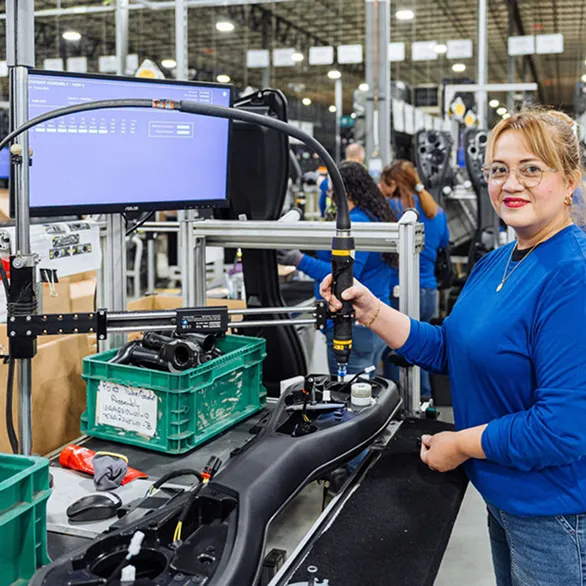 Worker holds powered torque tool above an assembly fixture with black plastic housing and green parts bin on workstation.