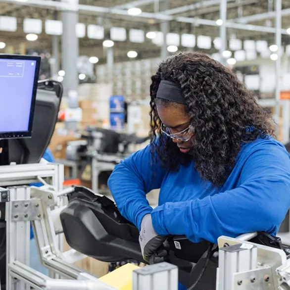 Worker in safety glasses leans over assembly fixture while securing internal components inside a black housing.