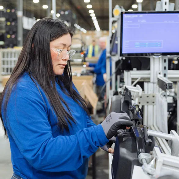 Worker wearing safety glasses installs a component into a black plastic assembly at a workstation with mounted display screen.