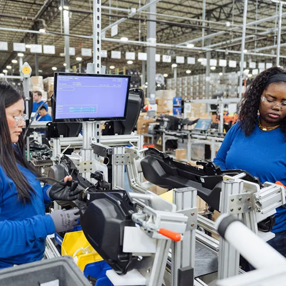 Worker assembles black plastic component at a production workstation with monitor and tooling in a warehouse facility.