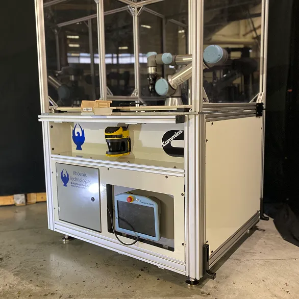 Angled view of enclosed robotic work cell showing collaborative robot arm, control unit with emergency stop button, and Phoenix Technologies and Cummins logos on the cabinet.