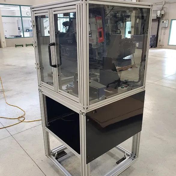 Angled view of an aluminum-framed enclosure with transparent panels and a black lower cabinet section inside a manufacturing facility.