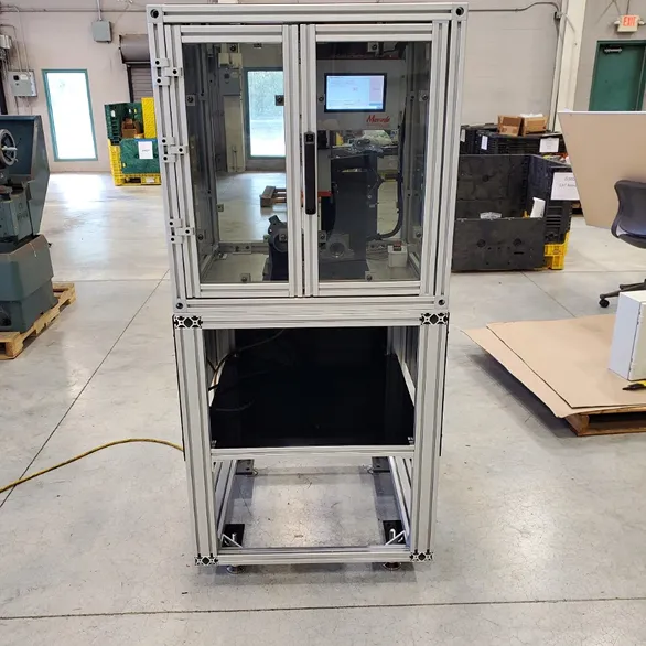 Enclosed aluminum frame machine with clear polycarbonate panels and a front access door, positioned on a factory floor.