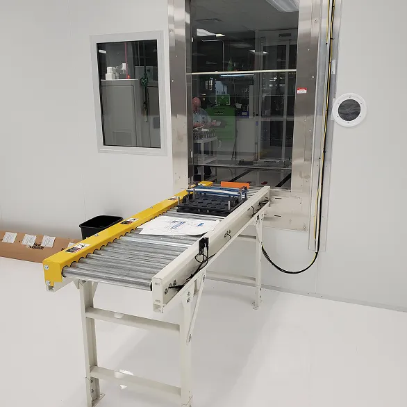 Roller conveyor extending through a stainless-steel wall pass-through with a fixture tray positioned on the conveyor surface.