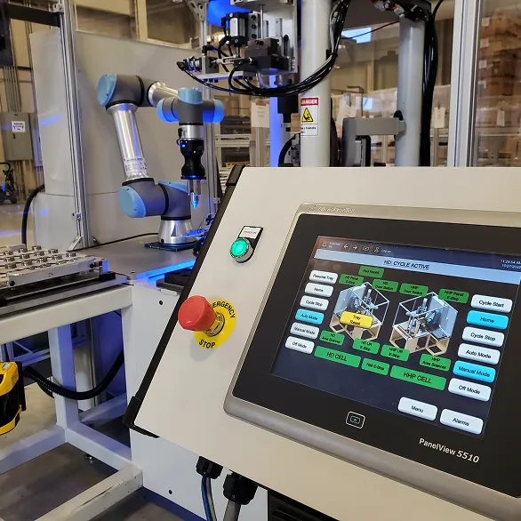 Control panel with a touchscreen interface and emergency stop button positioned in front of an enclosed automated cell containing a collaborative robotic arm and press equipment.