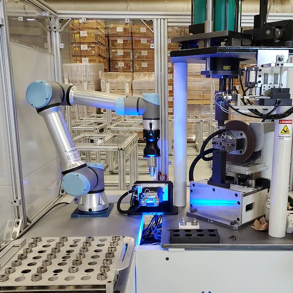 Enclosed automated workstation with a collaborative robotic arm positioned next to a vertical press and a fixture plate holding multiple metal parts inside a safety enclosure.