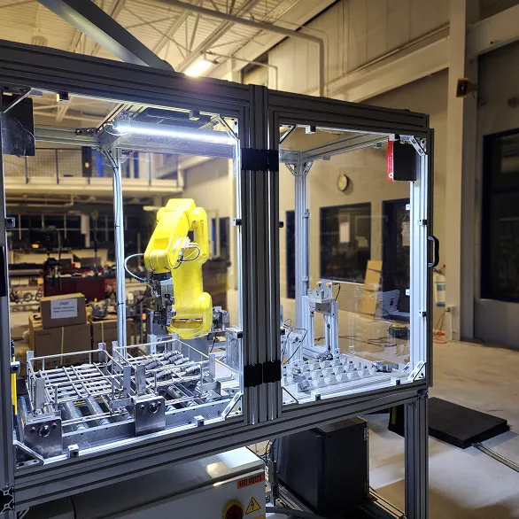 Front view of an enclosed robotic work cell with a yellow industrial robot loading or unloading parts from a fixture inside a transparent safety enclosure.