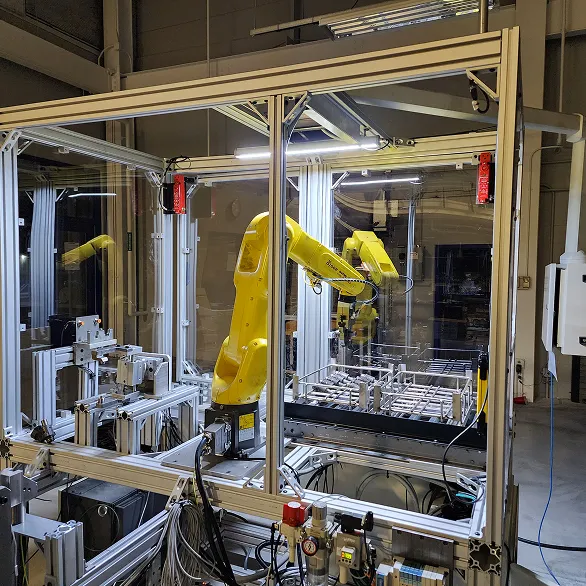Automated manufacturing cell with a yellow robotic arm, pneumatic components, and metal fixtures arranged inside an aluminum extrusion enclosure.