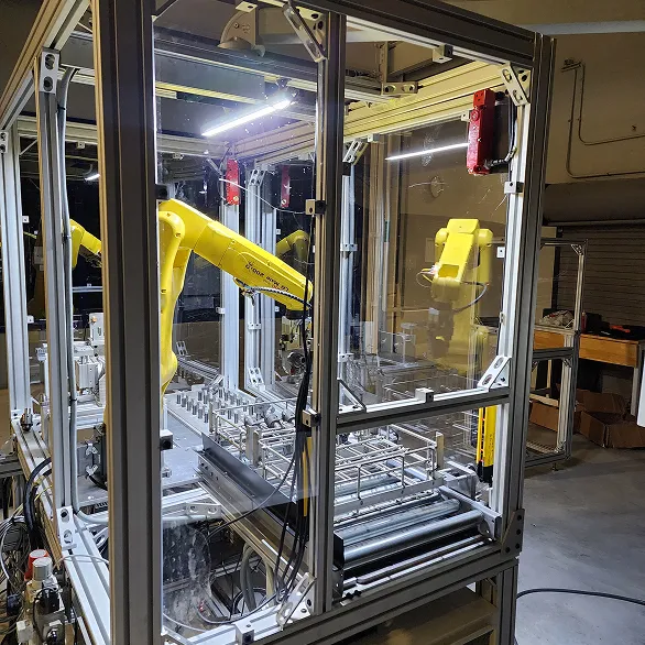 Side view of a guarded automation cell showing a yellow robotic arm, conveyor rollers, and part fixtures inside a clear safety enclosure on a factory floor.