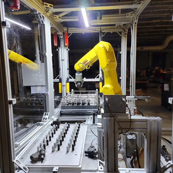 Enclosed automated cell with a yellow industrial robotic arm positioned above a fixture tray holding multiple metal components inside an aluminum frame enclosure.