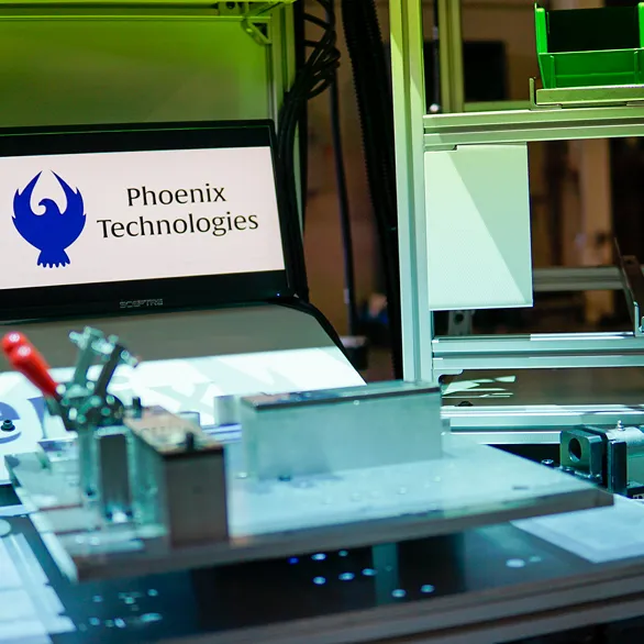 Close-up of workstation monitor displaying Phoenix Technologies logo beside an assembly fixture and handheld torque driver within an enclosed work cell.