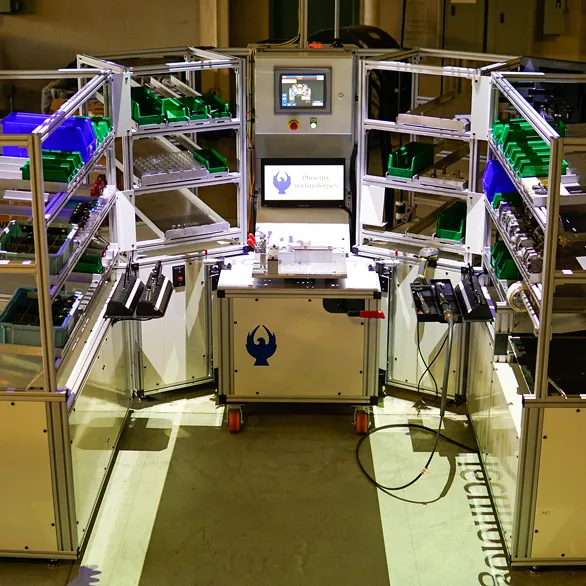 Automated work cell with aluminum framing, green parts bins, central control panel with touchscreen, and assembly fixture table inside a manufacturing lab. 