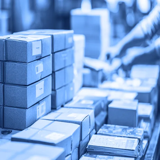 Stacks of small cardboard boxes and printed materials are arranged on a worktable in a warehouse, with a person in the background handling additional boxes.