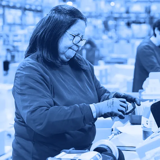 Worker wearing gloves applies tape to a cardboard box at a packing station with a monitor screen, bins of materials, and warehouse shelving in the background.