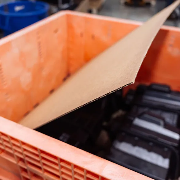 Flat cardboard liner positioned inside an orange plastic returnable tote containing black components.