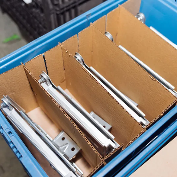 Metal components positioned in separate compartments formed by cardboard dividers inside a blue plastic returnable tote.
