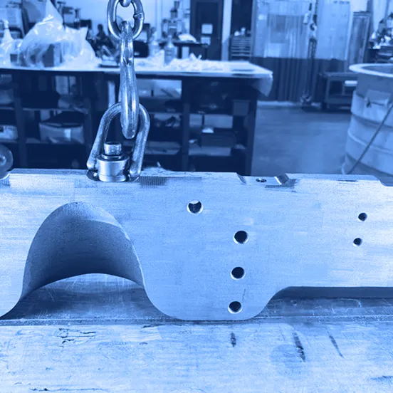 Large machined metal casting on a workbench with clean, uniform metal surfaces and drilled holes visible, showing removal of rust, with lifting hardware attached.