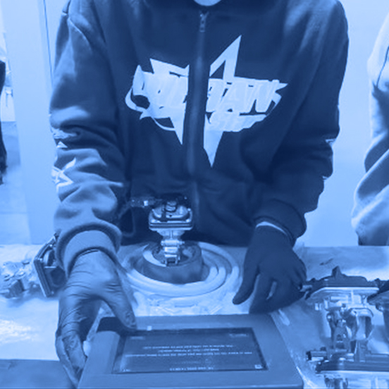 Two workers wearing gloves assemble metal components at a workbench with a tablet screen and tools visible in front of them.