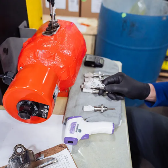 Worker’s gloved hand holding a metal sensor near a heated testing device on a worktable.