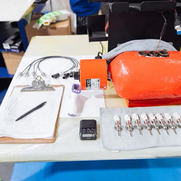 Worker seated at a worktable reviewing a small component next to testing equipment, paperwork, and handheld measurement tools.