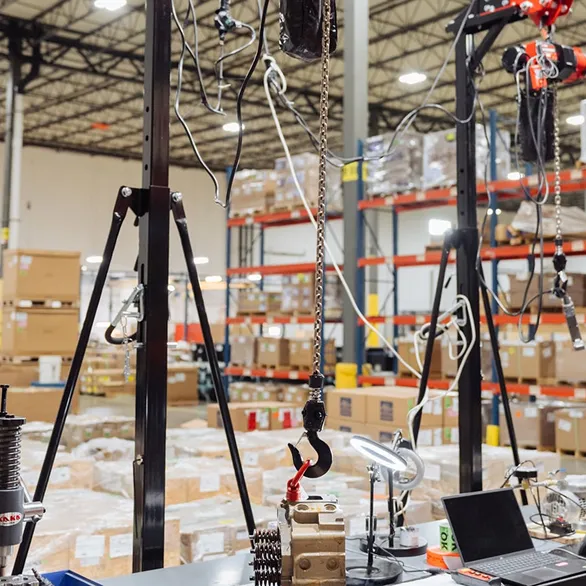 Overhead hoists with chain hooks positioned above metal cylinder heads on an inspection workbench inside a warehouse.