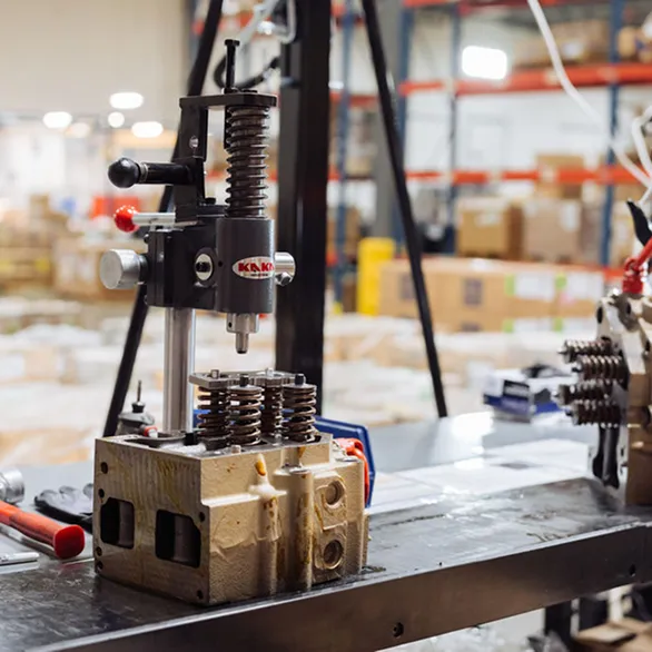 Close-up of a metal cylinder head secured on a worktable beneath a manual valve spring compressor with hand tools nearby.