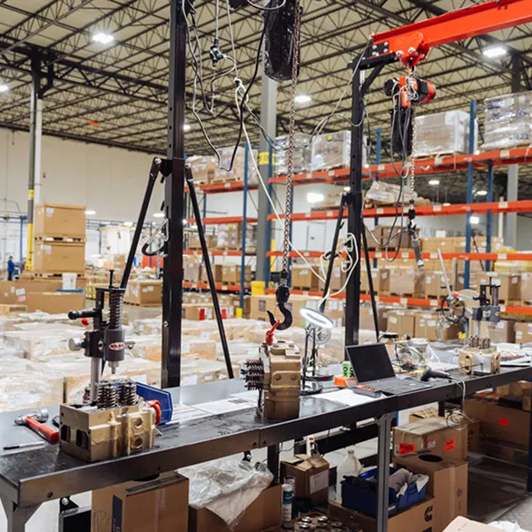 Industrial workbench with multiple metal cylinder heads positioned under manual valve spring tools and overhead hoists in a warehouse setting.