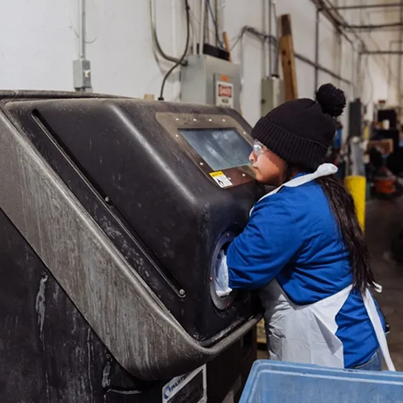 Worker wearing gloves and a protective apron reaches into a sealed cabinet-style machine used for part cleaning or testing.