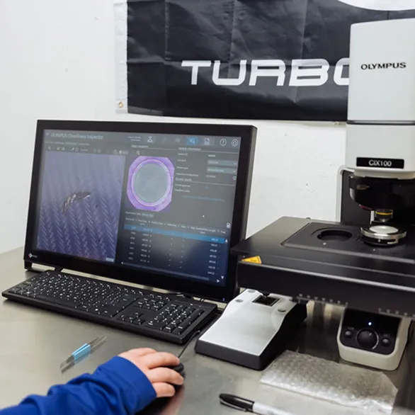 Computer monitor and microscope on a metal workbench display a magnified circular image used for part inspection.