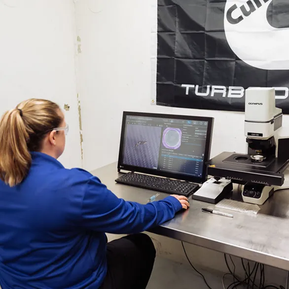 Worker seated at a stainless steel table operates a microscope connected to a computer displaying a circular inspection image.