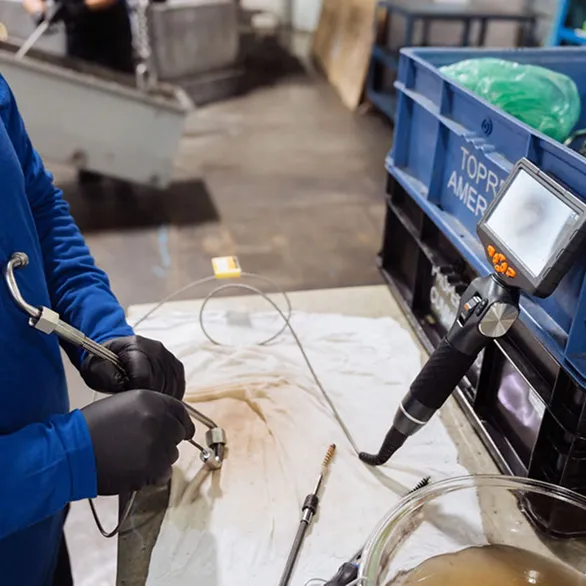 Close-up of gloved hands holding a flexible inspection probe and metal fitting on a stained work surface beside a digital borescope monitor.