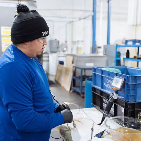 Worker wearing gloves and safety glasses uses a handheld borescope probe at a metal worktable next to plastic bins and inspection tools.